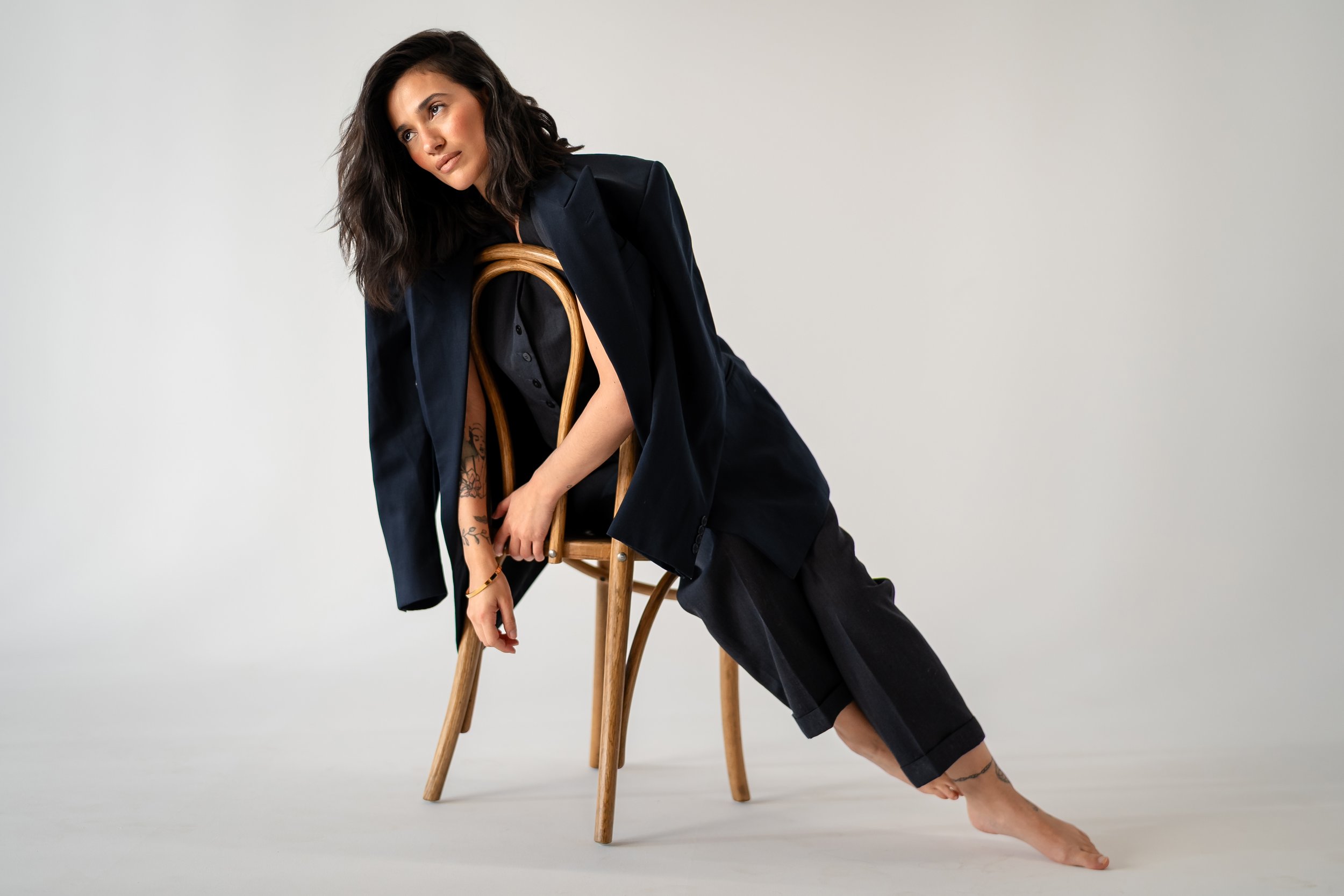 Woman with dark hair wearing a black coat and pants, leaning on a wooden chair in a studio with a plain background.