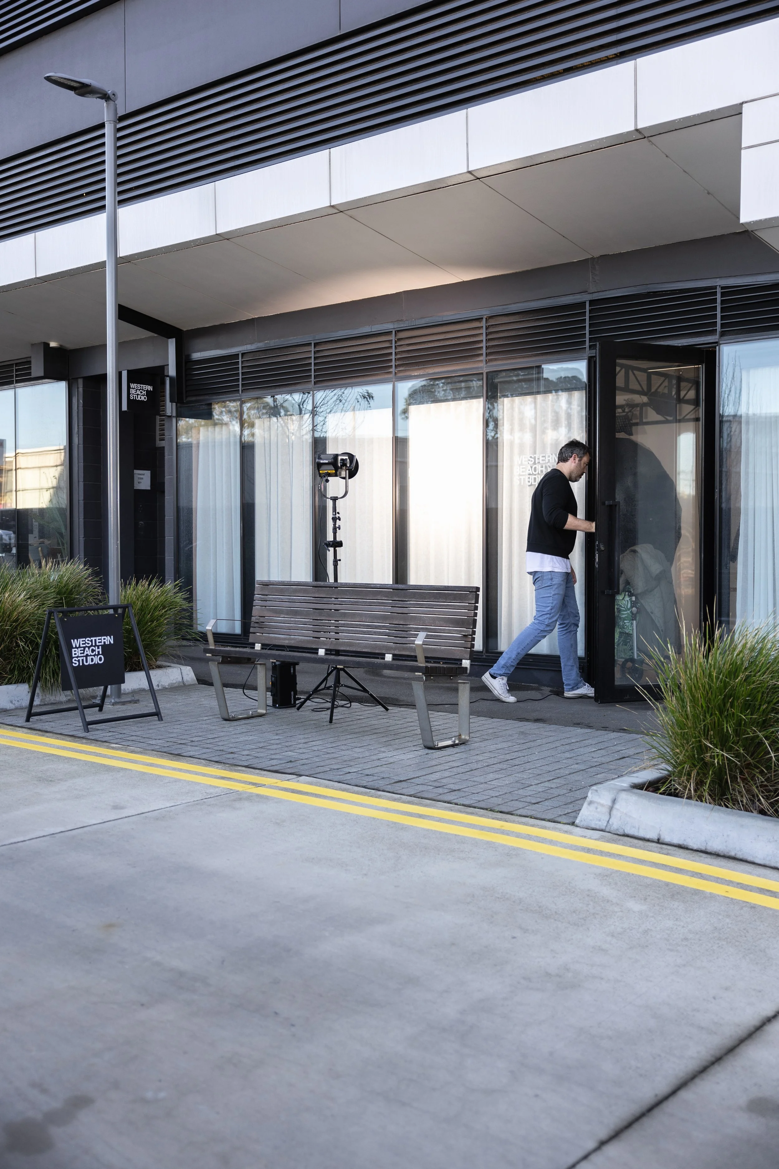 A man entering a building called Western Beach Studio, with a bench and sign outside, and film equipment nearby.