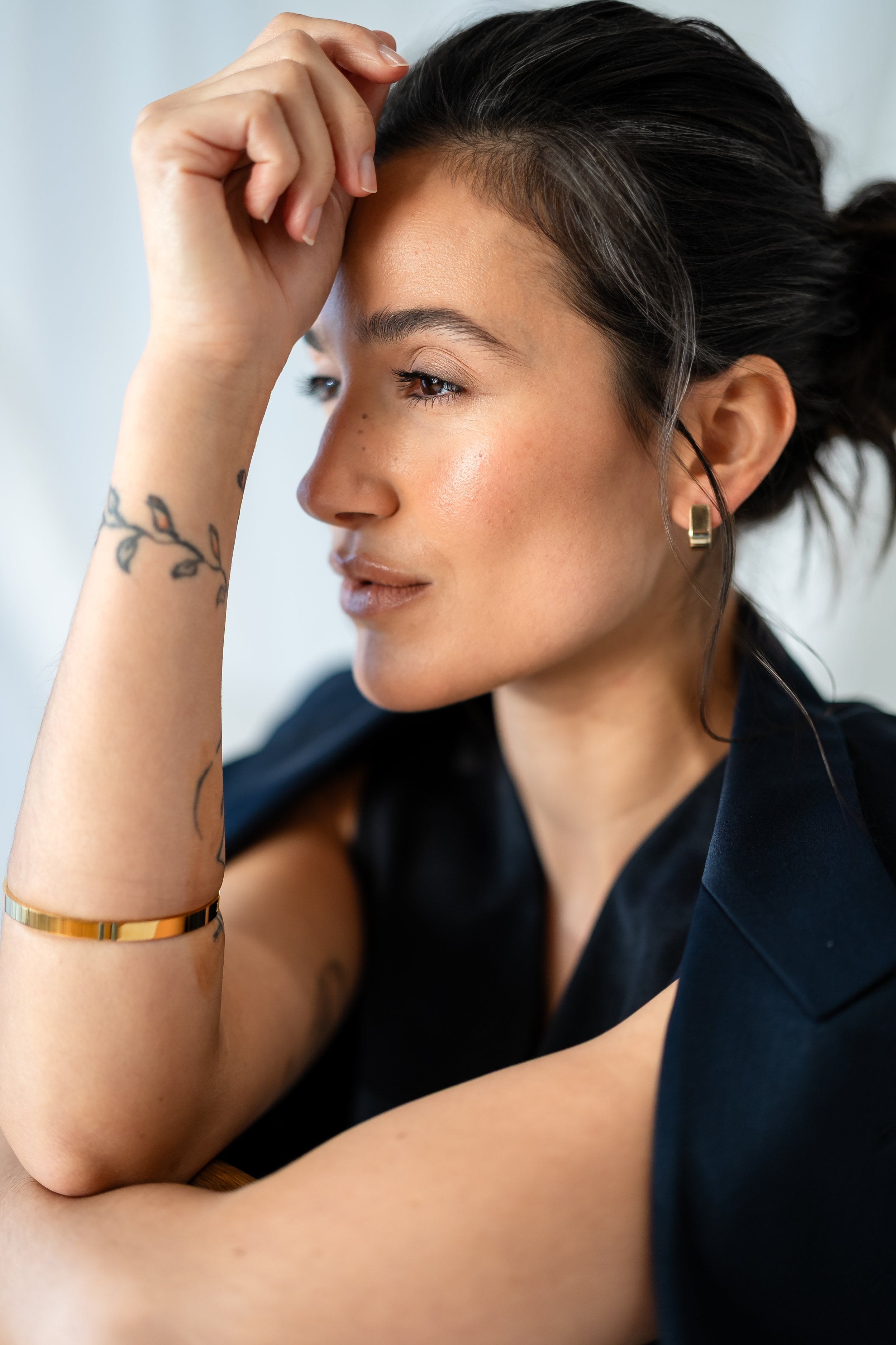A woman with dark hair styled in a loose ponytail, wearing hoop earrings and a black top, resting her chin on her hand while looking thoughtful.