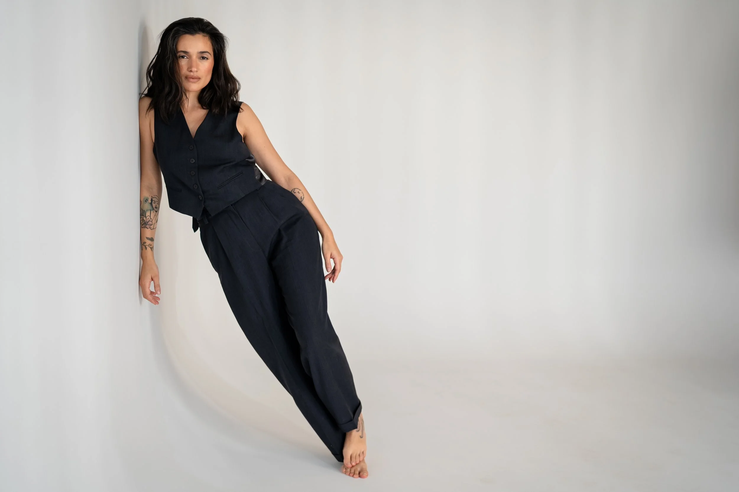 Woman with dark hair and tattoos wearing a black sleeveless vest and loose pants, standing against a white background.
