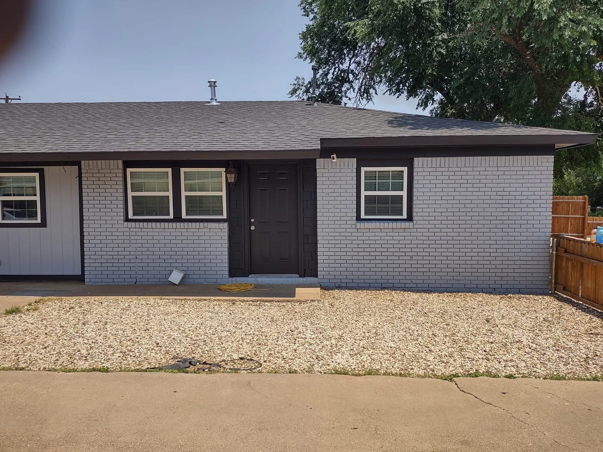 A single-story house with white painted brick exterior, dark gray roof, and dark door. The house features three windows with white trim and dark shutters, and a wooden fence on the right side. The front yard has gravel and concrete pavement, with a yellow extension cord and a small white object near the house.