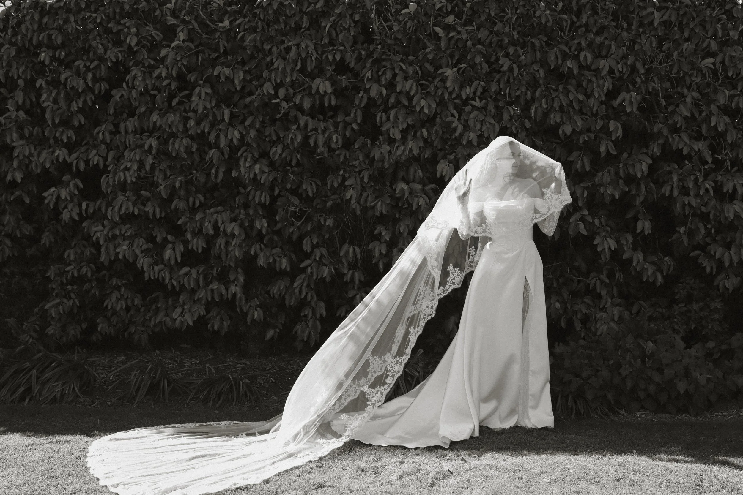 A woman in a wedding dress with a long train and lace veil stands outdoors in front of a hedge.