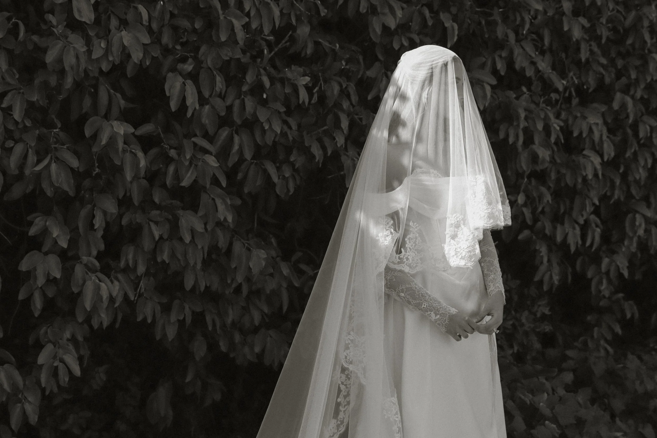 Black and white photo of a bride in a wedding dress with a veil, standing against a dark leafy background.