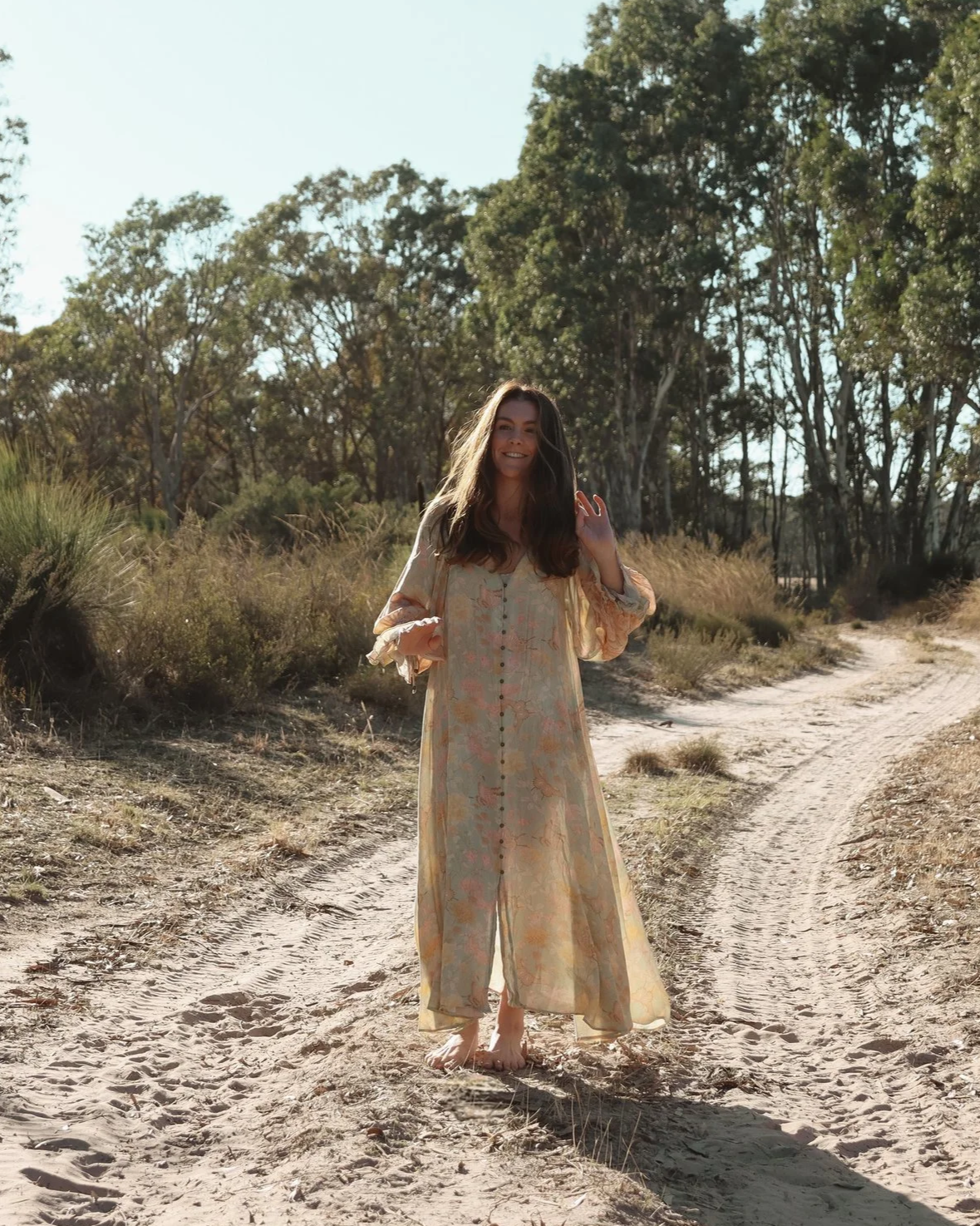 A woman with long brown hair smiling and waving, walking barefoot on a dirt path in a natural outdoor setting with trees and bushes.