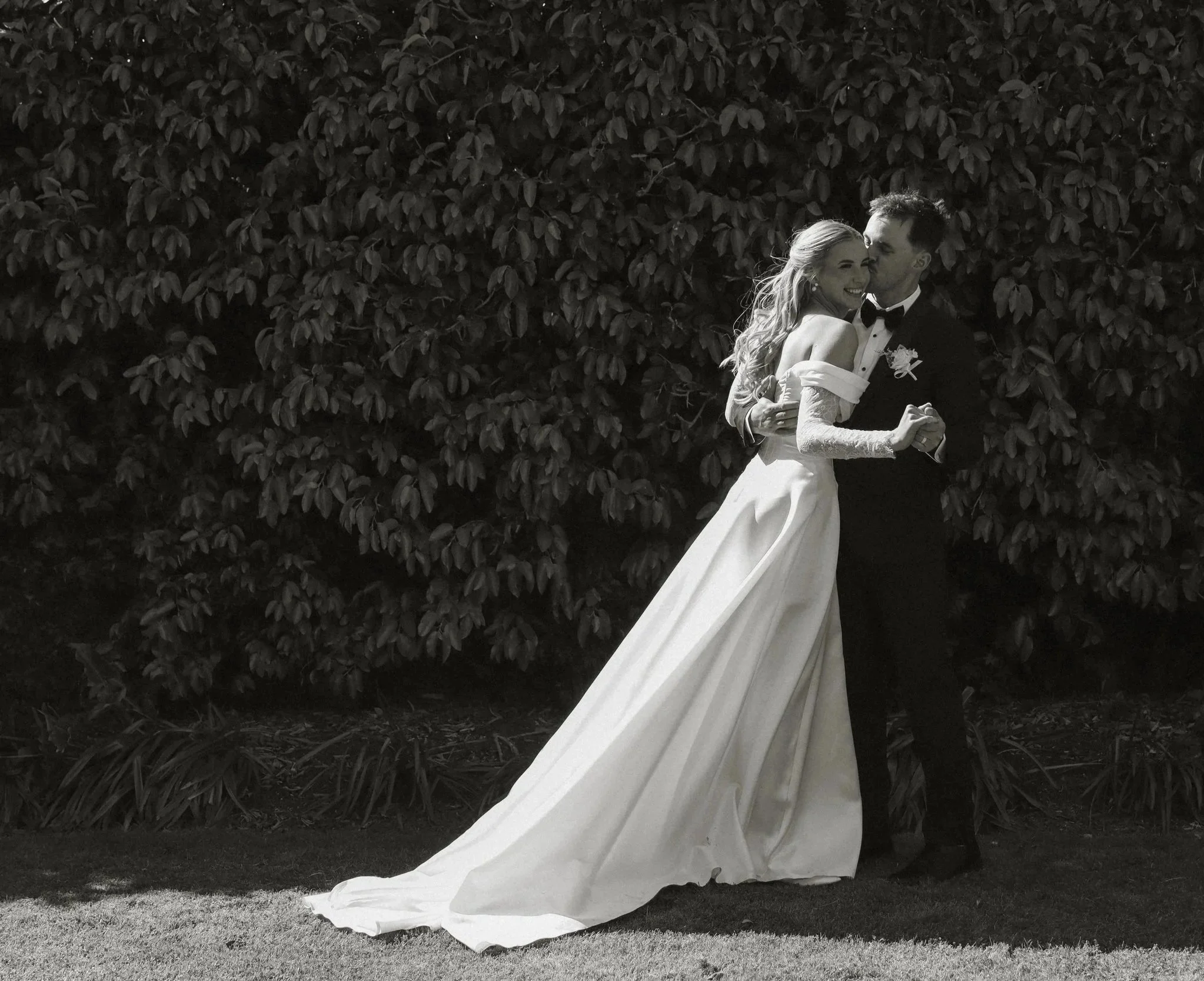 A bride and groom dance outdoors in front of a dense leafy hedge, the bride wearing a long white dress and the groom in a tuxedo, sharing a joyful moment.