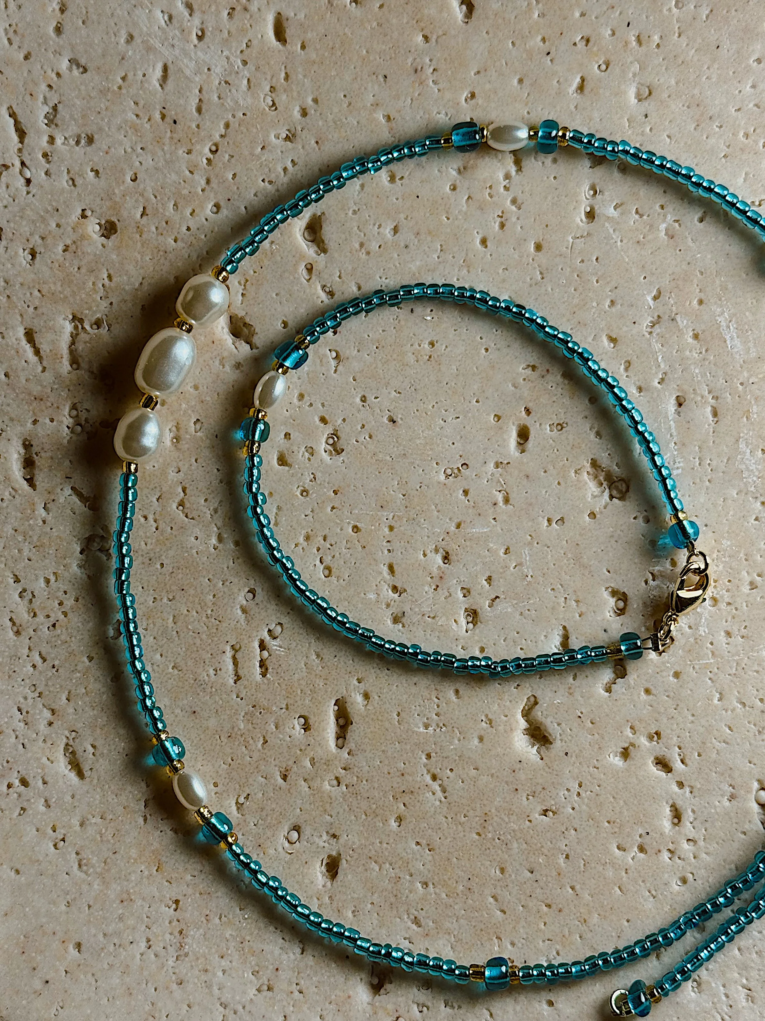 Blue beaded jewelry, including a necklace and bracelet, with some pearl accents, on a beige textured surface.