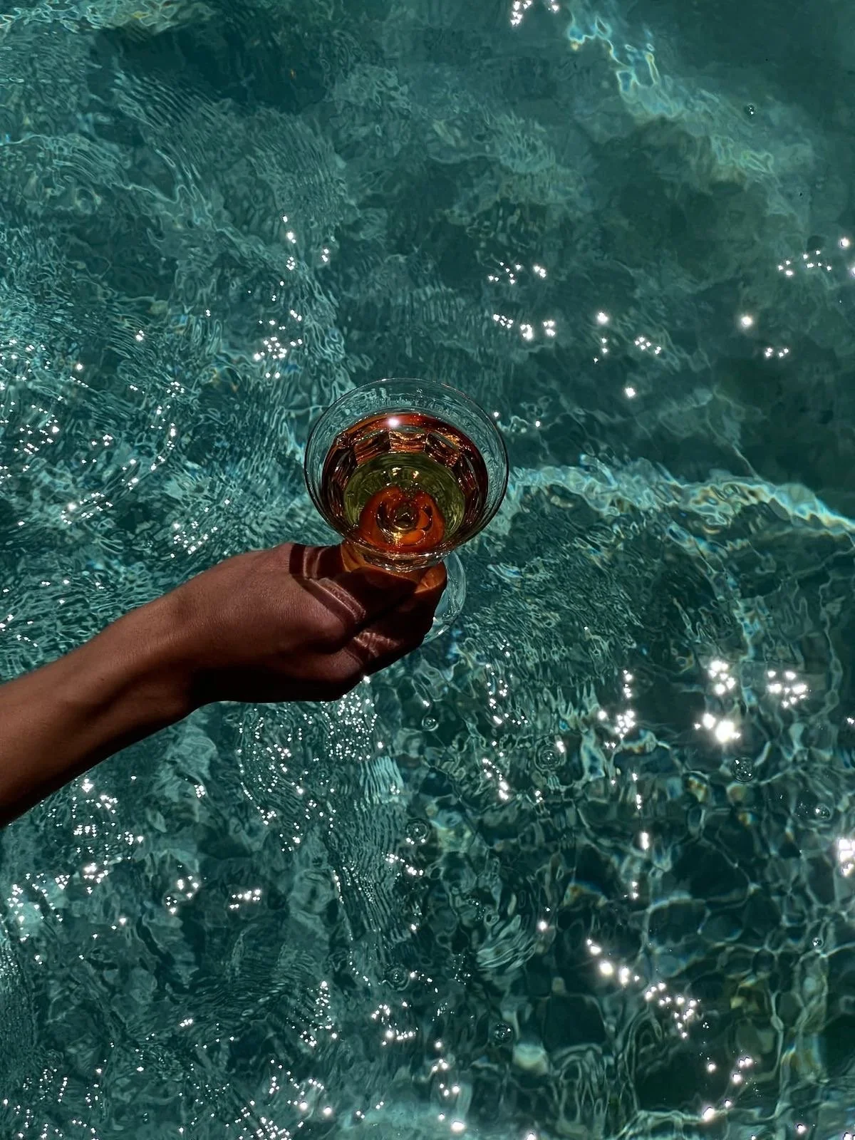 A person holding a cocktail glass with amber-colored liquid over a swimming pool with turquoise water and sparkling reflections.