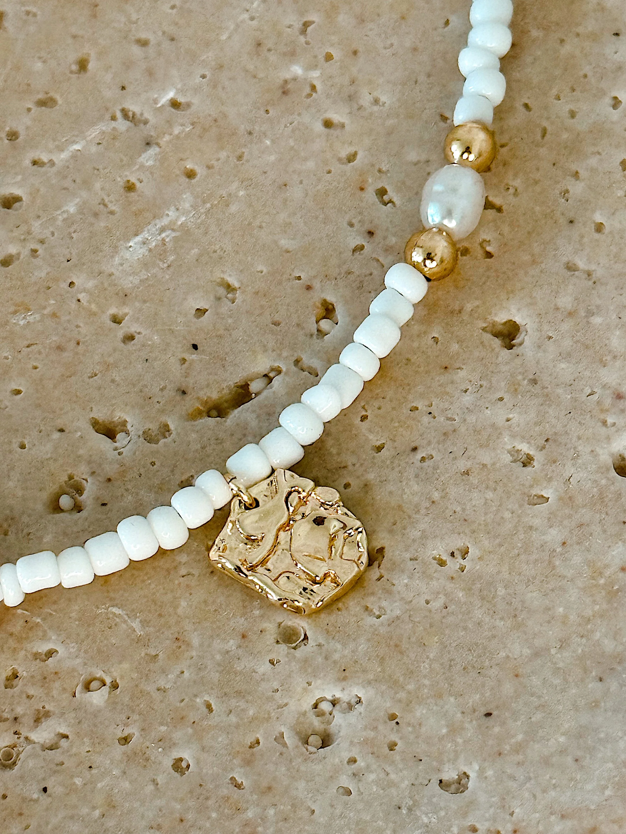 Close-up of a necklace with small white beads, larger gold and white beads, and a gold pendant on a speckled beige surface.