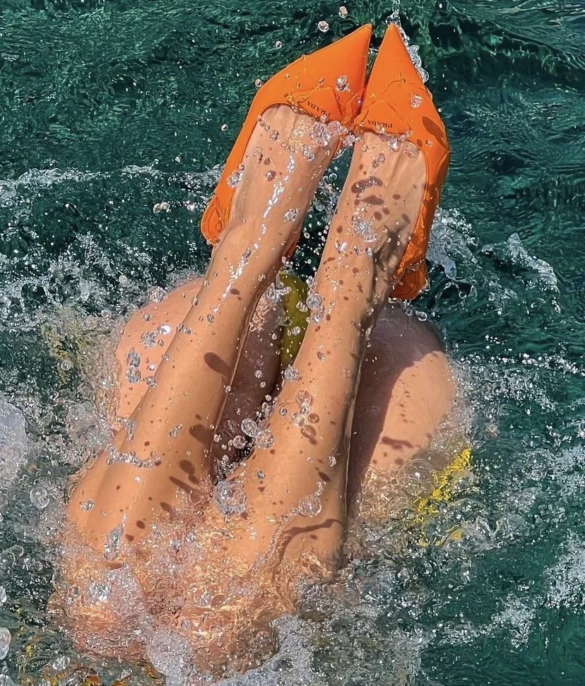 Person wearing orange water shoes swimming underwater, with legs extended upward and splashing water.