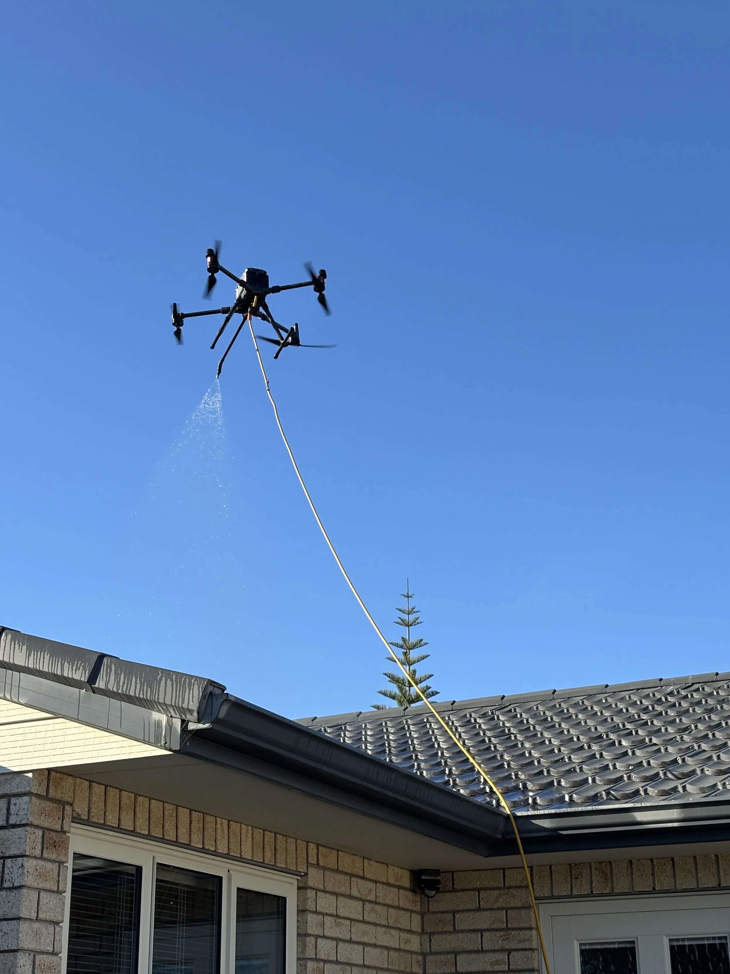 Drone spraying water or a liquid over a house roof against a clear blue sky.