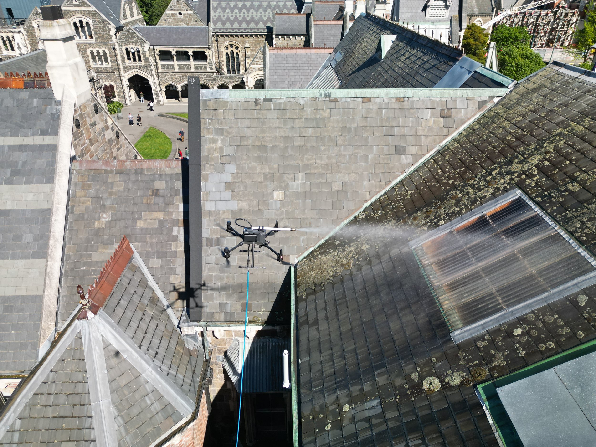 View of rooftops in a historic area with an agricultural drone spraying water or pesticides on a roof. The rooftops are covered with slate tiles, some showing moss or lichen. In the background, there is a stone building with arched windows and an entrance, and small groups of people walking on a courtyard.