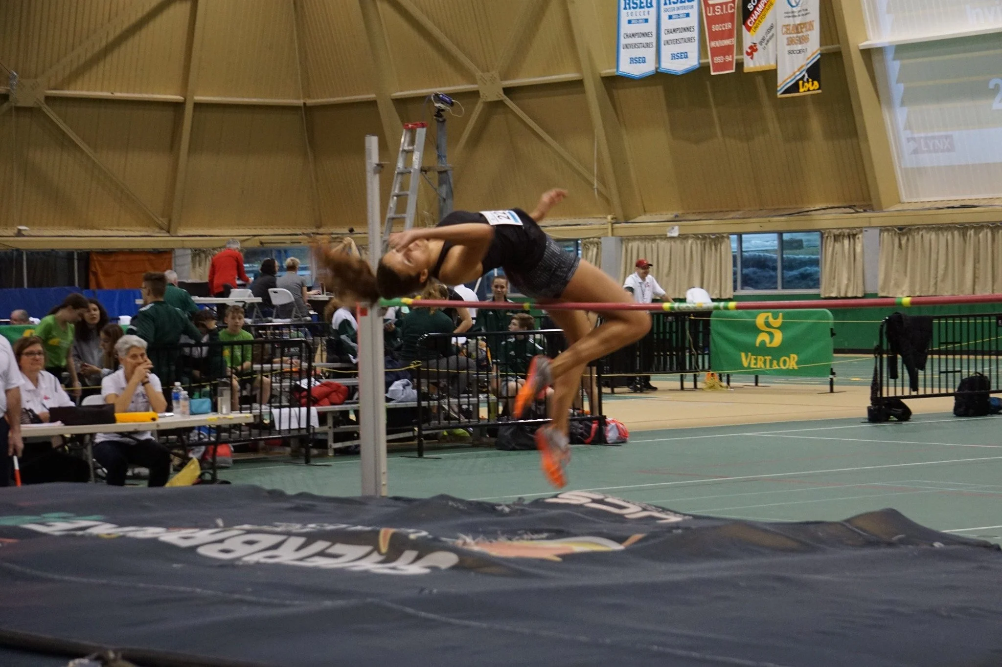 Une athlète en train de sauter par-dessus la barre lors d'une compétition d'athlétisme en intérieur, avec un public et des juges en arrière-plan.