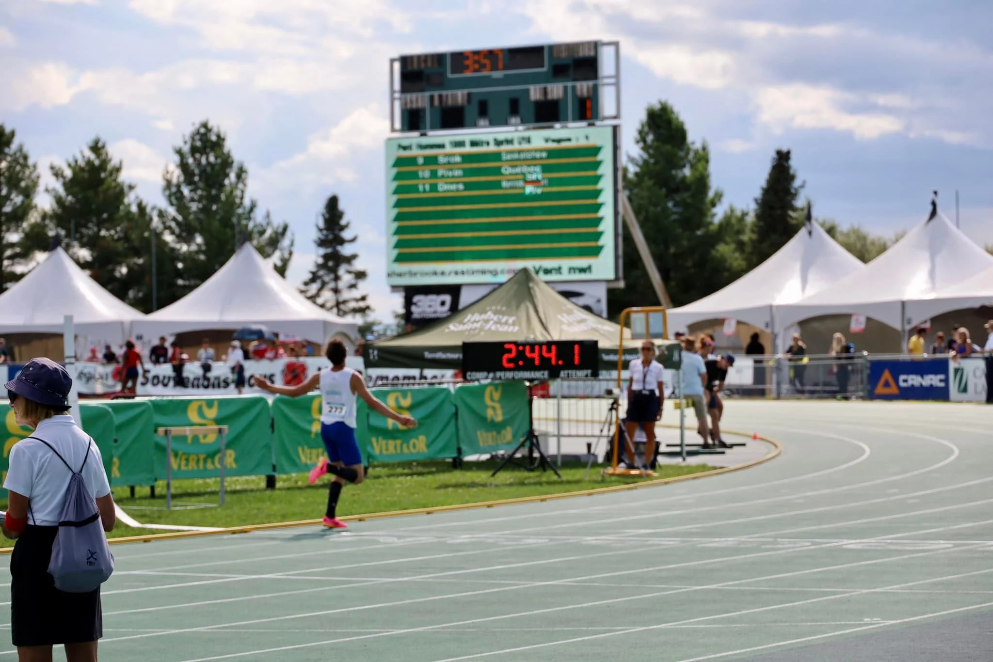 Un athlète franchit la ligne d'arrivée lors d'une compétition de course sur piste, avec un tableau d'affichage numérique montrant 2:44.1, et des tentes blanches en arrière-plan.