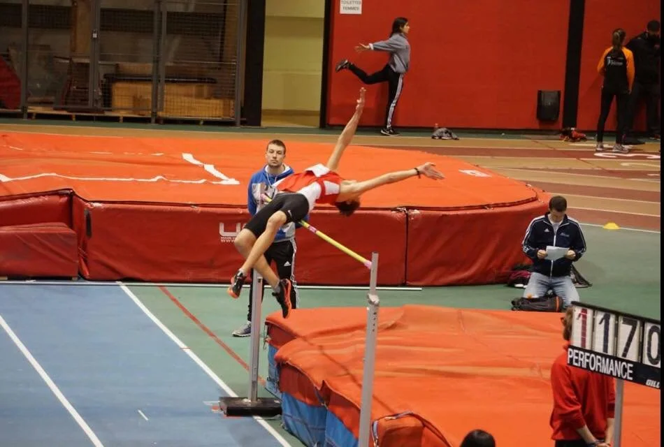 Athlète en plein saut lors d'une compétition de saut en hauteur dans une salle d'athlétisme.