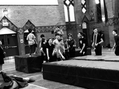 Groupe de jeunes en train de faire des exercices ou une activité dans une salle d'école ou de gym, avec des fenêtres en vitrail en arrière-plan.