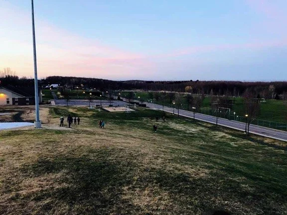 Un parc avec des personnes se promenant, une piste cyclable, des lampadaires et un ciel au crépuscule.