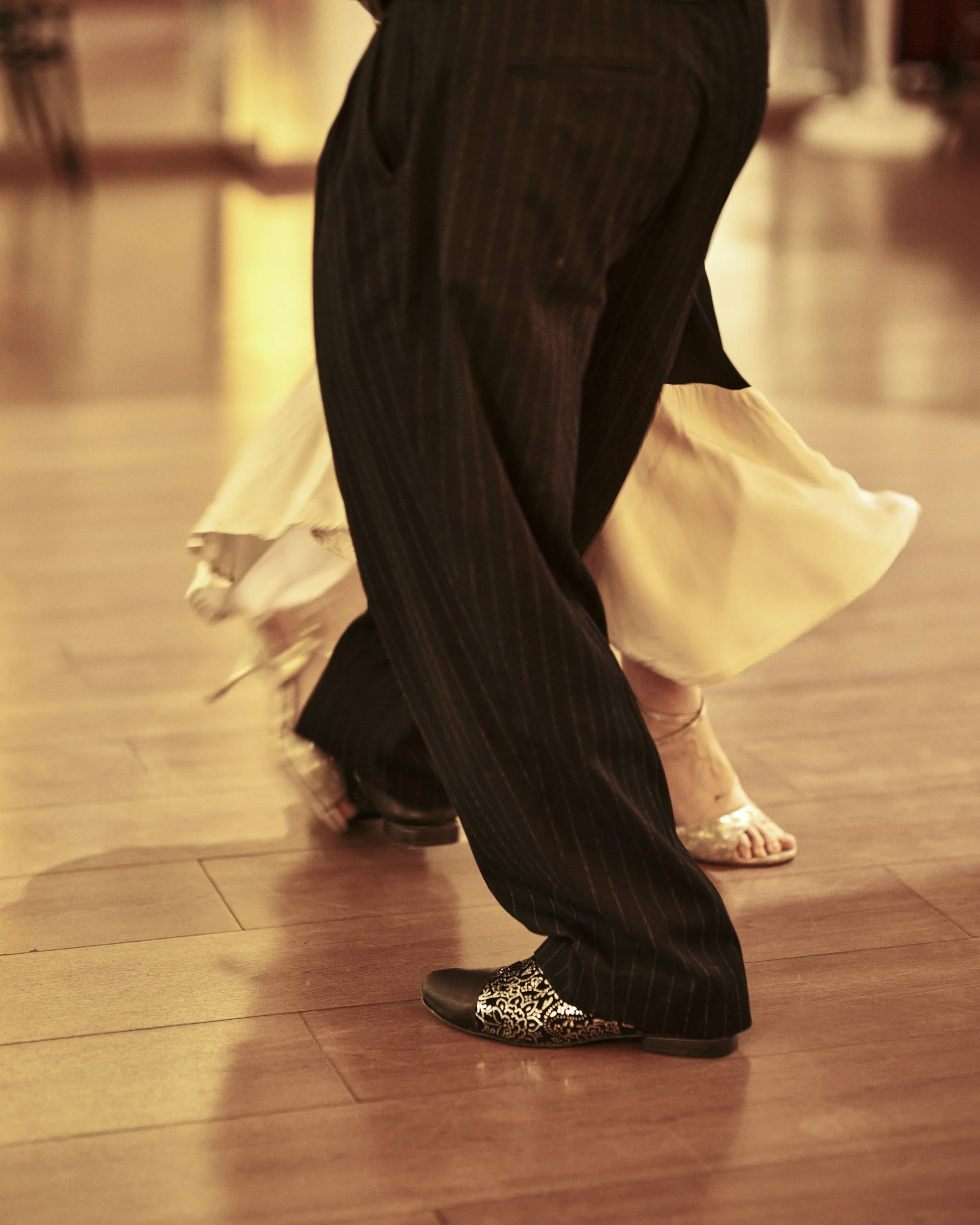 Close-up of the lower halves of two people dancing, one wearing black pinstripe pants with decorative shoes, and the other in a flowing cream-colored dress and heels, on a wooden floor.