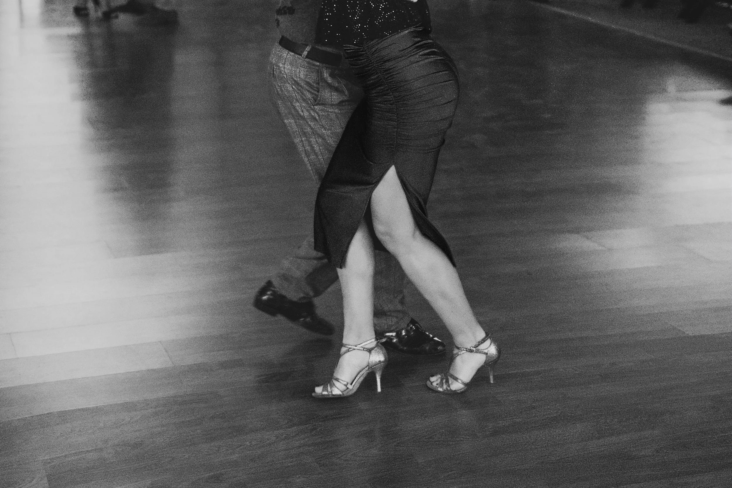 Couple dancing closely on a wooden floor, focusing on their legs and feet, with the woman's dress revealing her legs and wearing high heels.