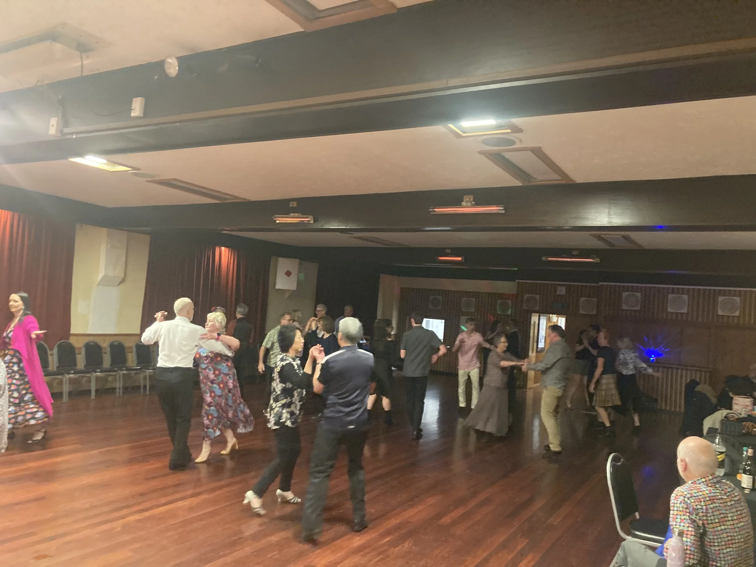 People dancing in a dimly lit banquet hall with wooden floors, some seated at tables, and a couple of wall-mounted speakers visible.