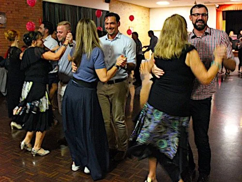 People dancing in pairs at a social event in a hall with decorations.