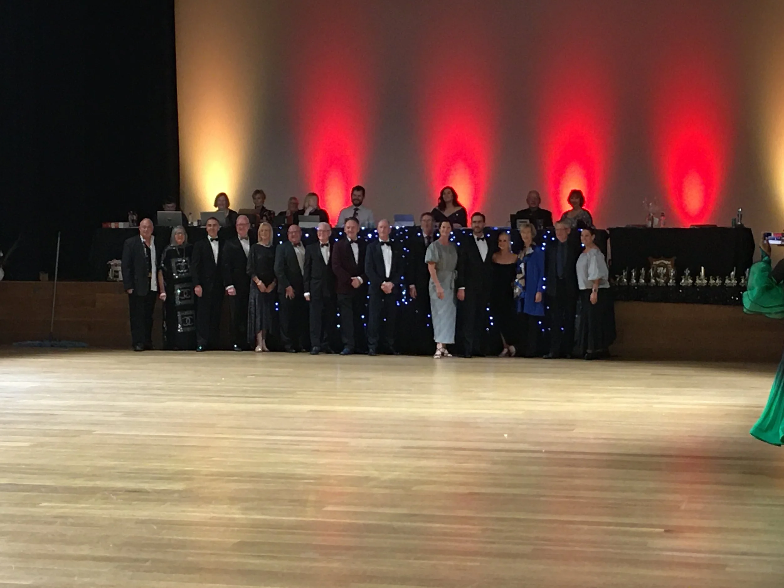 A group of people dressed in formal attire standing on a stage with a decorated background. Behind them, there are people working at laptops and sound equipment, with stage lighting creating red and yellow spotlight effects on the wall.
