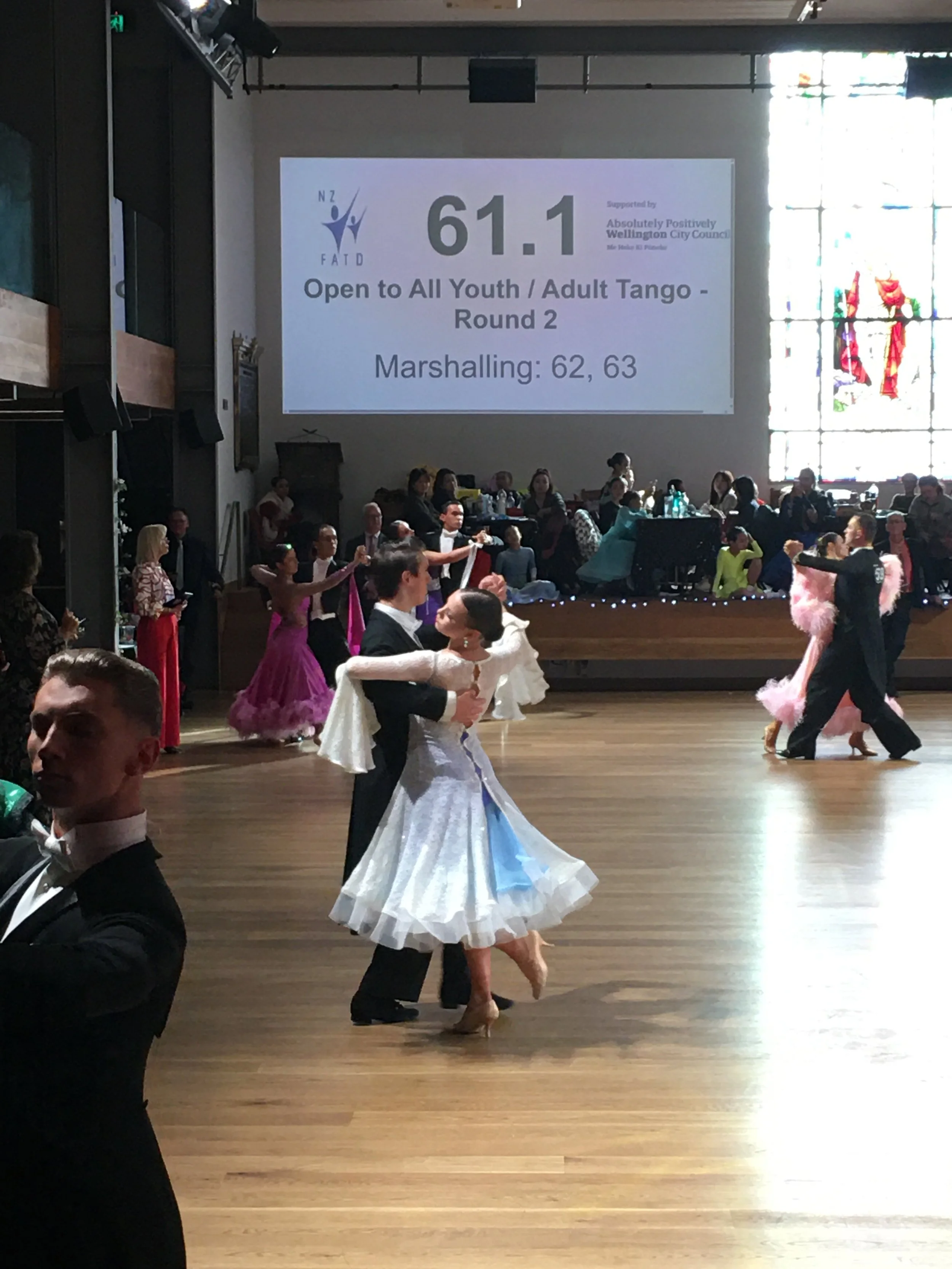 Couples in ballroom dance costumes performing tango in a large hall with a wooden floor. Audience seated along the side, and a large projected screen displaying competition details in the background.