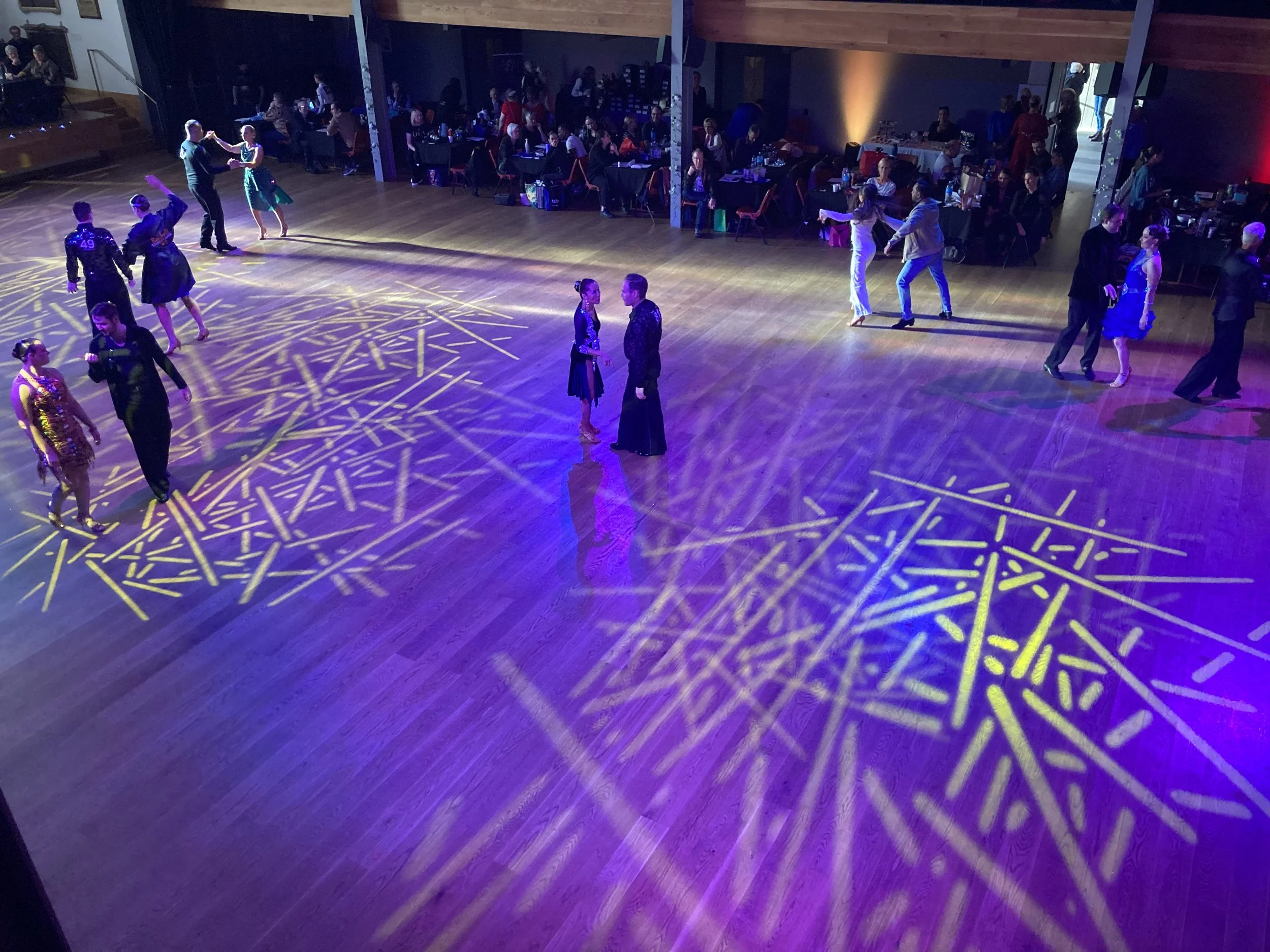 Couples dancing on a wooden floor illuminated by purple and yellow stage lights, with seated guests at tables watching.
