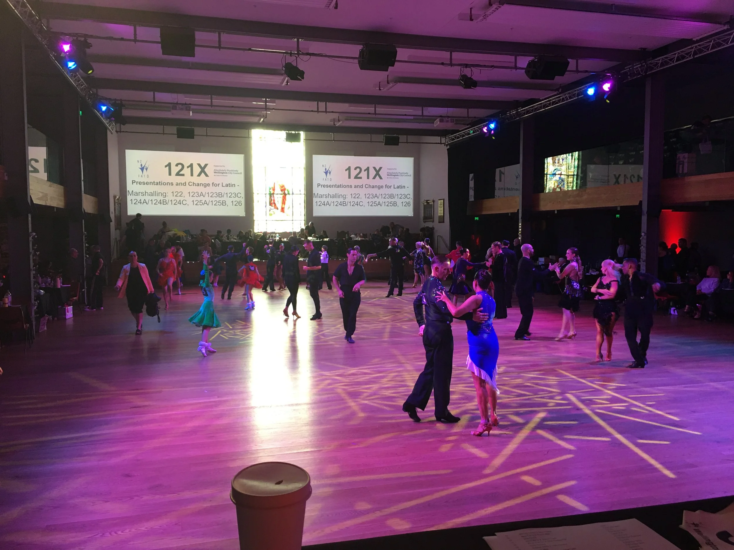 Dance event in a large hall with couples dancing on the floor under purple and pink lighting, with large screens displaying '121X' and text about presentations and change for Latin dance styles.