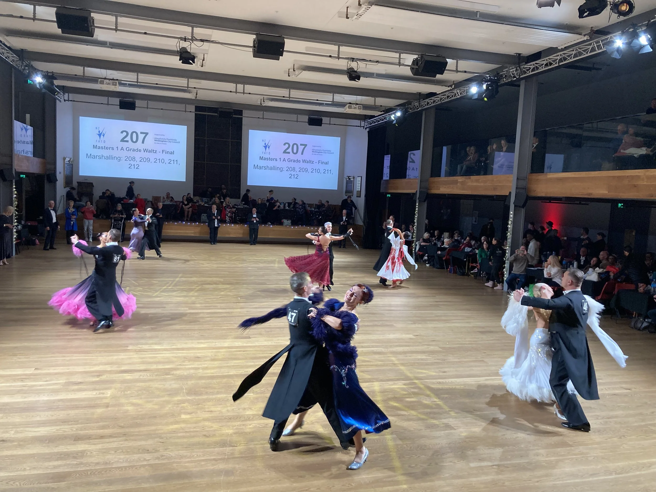 Couples ballroom dancing in formal attire during a dance competition on a wooden floor, with audience members watching from the sides and large screens displaying event information.