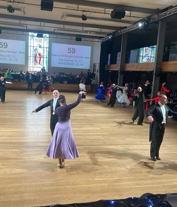 Couples dancing at a ballroom dance competition, with some couples in elegant gowns and tuxedos. The dance floor is wooden, and there is a large screen displaying competition information in the background.