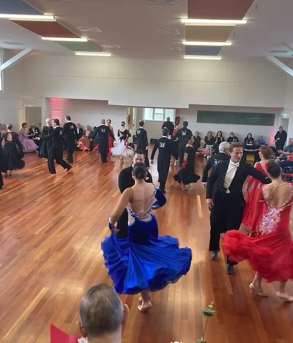 People dancing in formal attire in a spacious dance hall with wooden floors, white walls, and a blackboard.