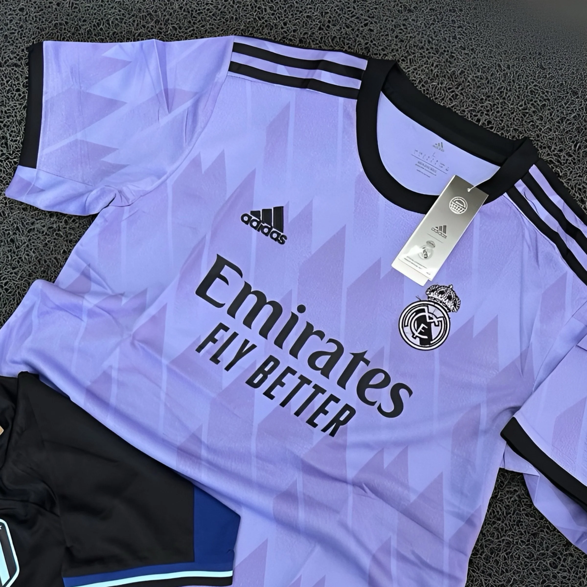 A lavender Real Madrid soccer jersey with black accents, Adidas logo, and the 'Emirates Fly Better' sponsor, lying flat on a textured black surface.