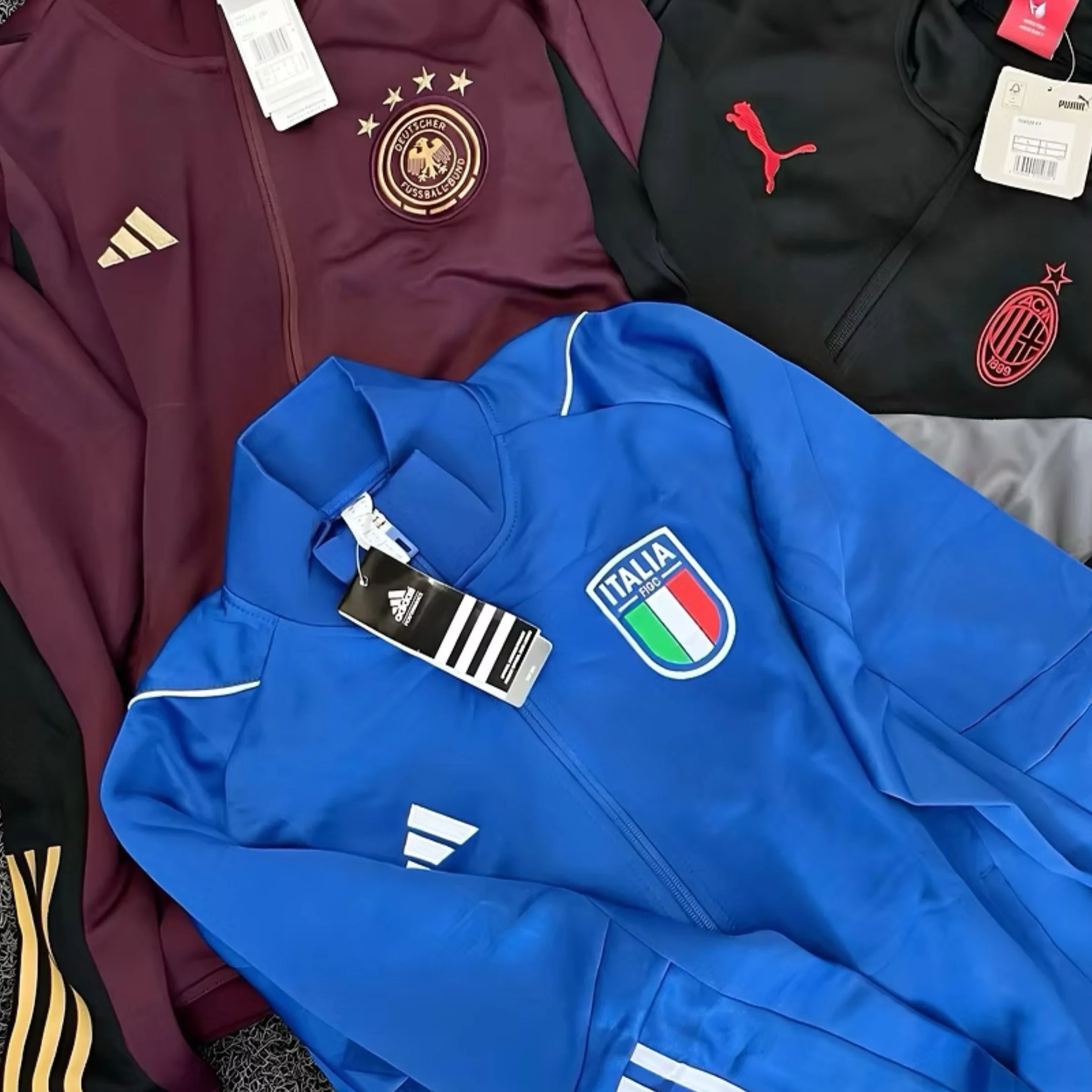 Three sports jackets lay on a surface. The left jacket is maroon with a gold logo of the German Football Association. The right jacket is black with pink Puma and AC Milan logos. The bottom jacket is blue with the Italian national football team crest and Adidas logo.