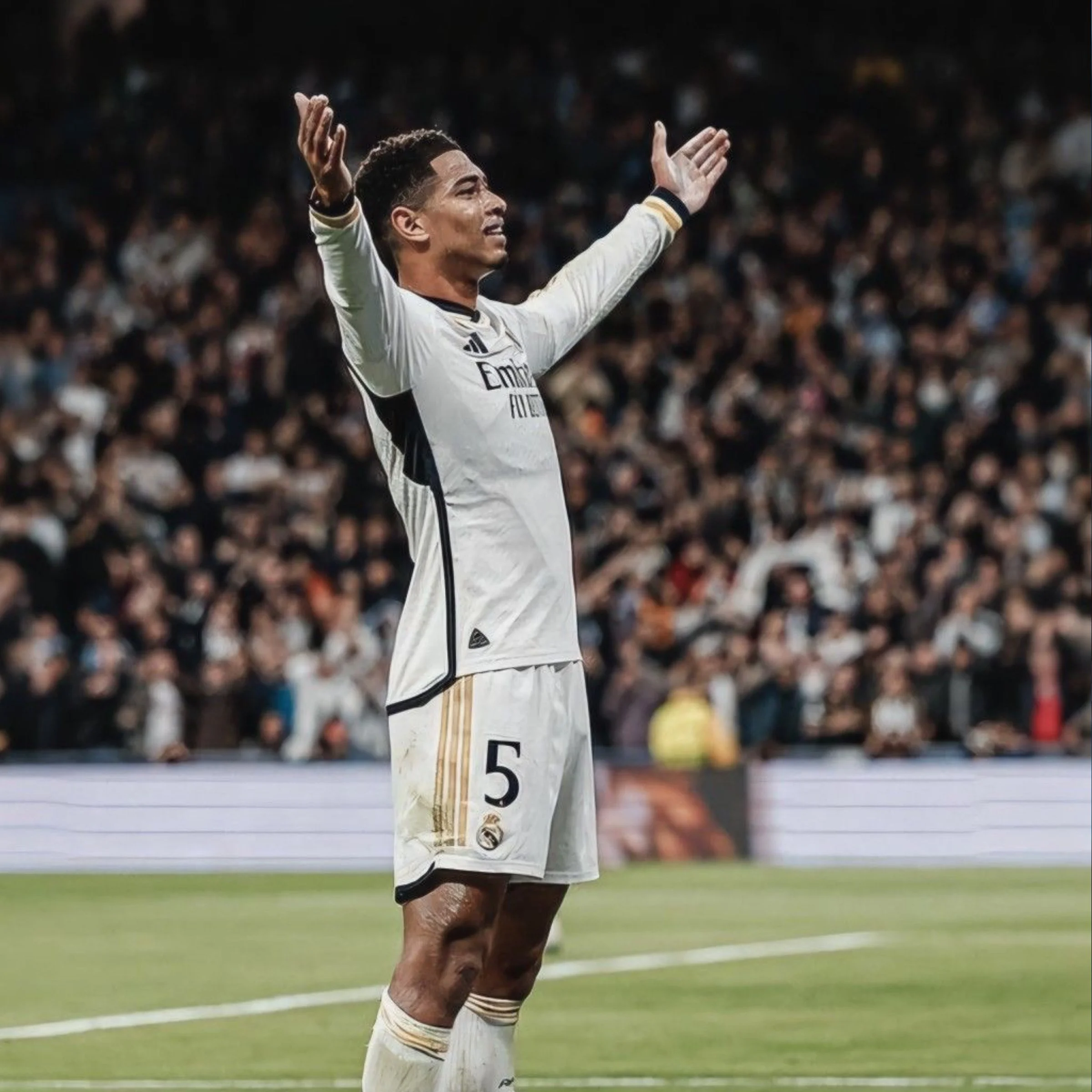 A soccer player wearing a white uniform with the number 5 and gold stripes raises his arms in celebration on the field with a blurred crowd in the background.