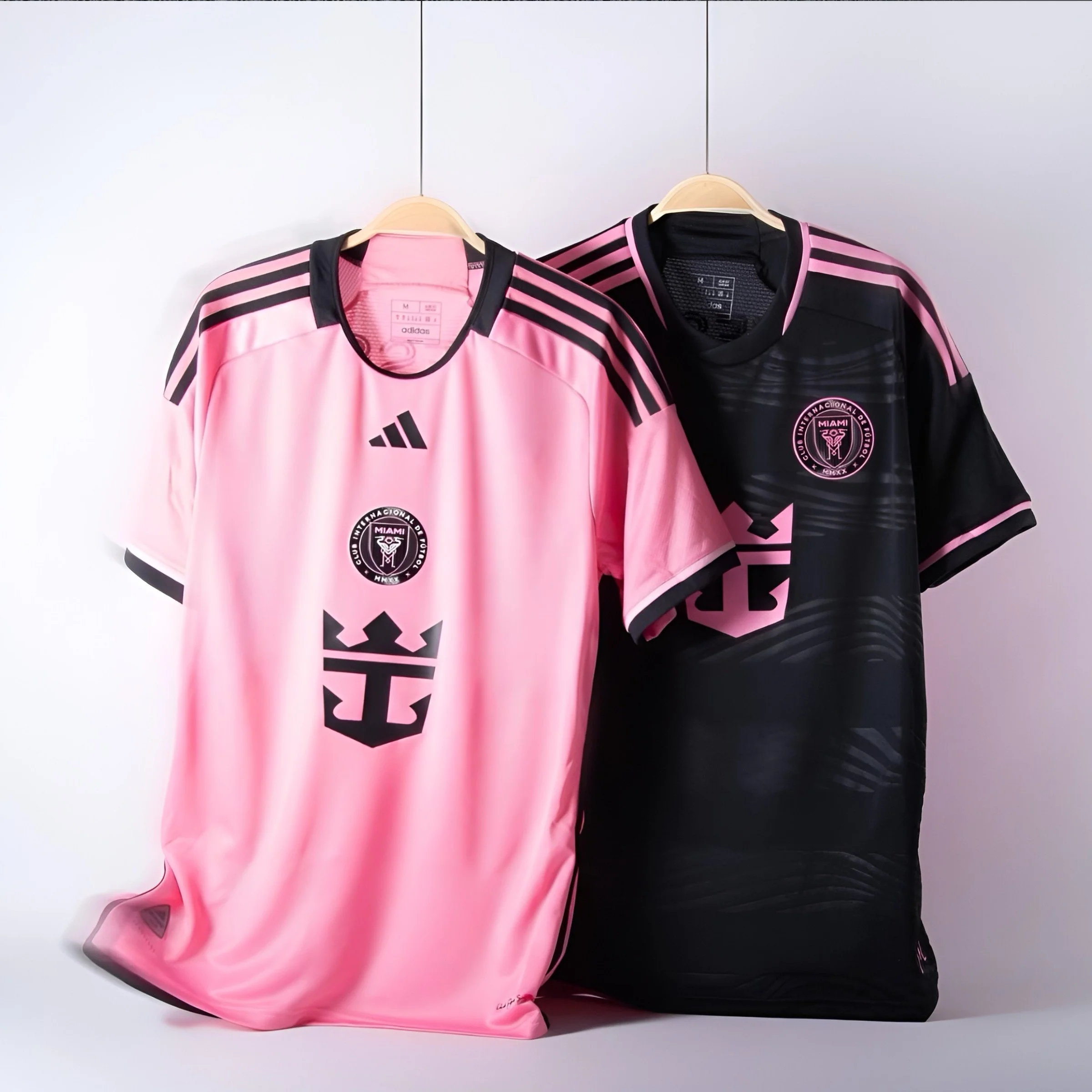 Two Miami soccer jerseys, one pink and one black with pink accents, hanging on hangers against a plain wall.