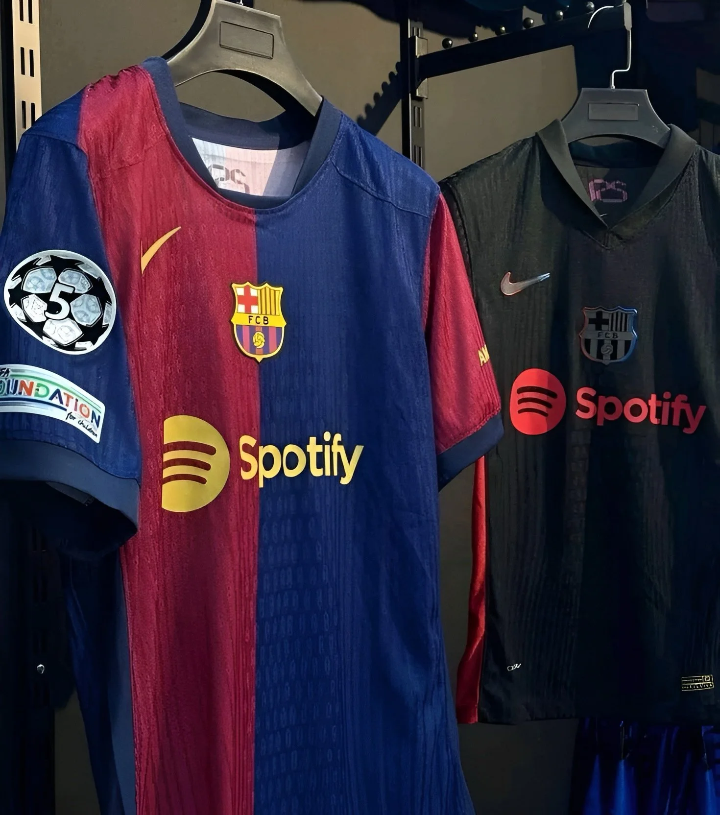 Two FC Barcelona soccer jerseys hanging on black hangers on a rack, featuring sponsor logos including Spotify. One jersey is in red and blue with the club crest and a sleeve patch, the other is black with the crest and sponsor logo.