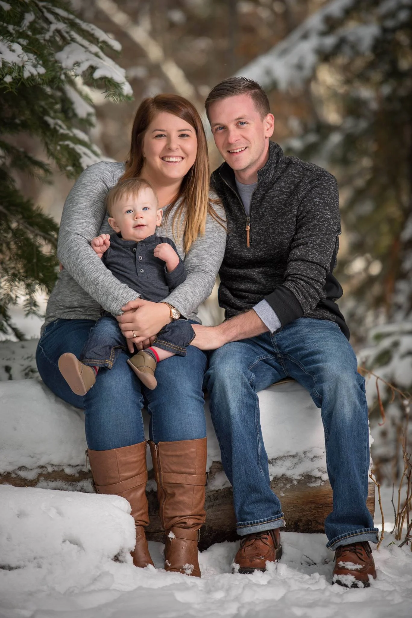 A family of three sitting on a snow-covered log outdoors in winter. The woman is holding a young child on her lap, and the man is sitting beside her. They are all smiling, with snow-covered trees in the background.