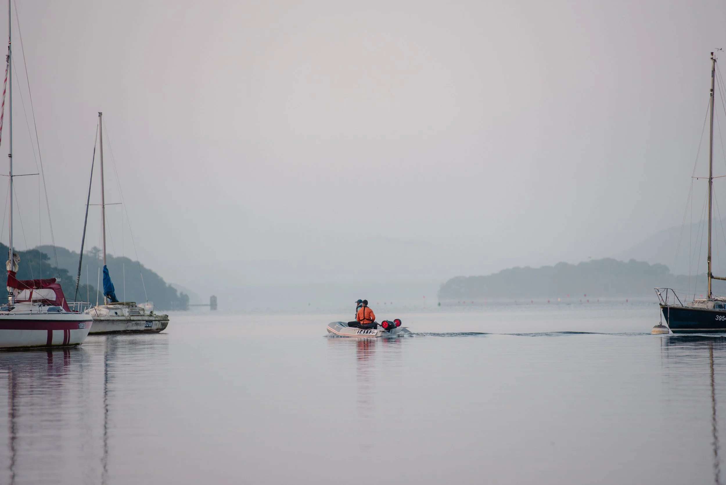 A person in an orange life jacket riding a small motorboat on calm water, with sailboats docked on either side, and distant islands in the background, under a hazy sky.