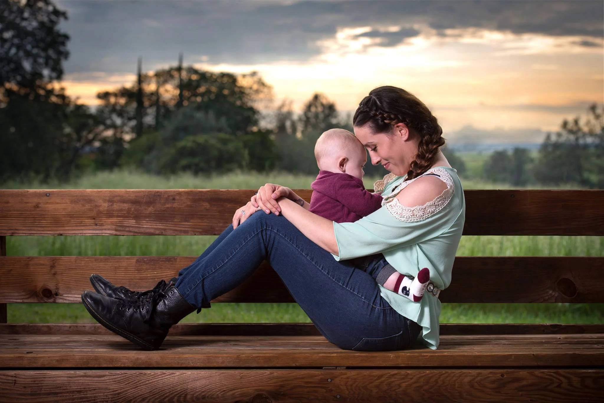 A woman and a young child sharing an intimate moment on a wooden park bench during sunset, with a scenic background of trees and a cloudy sky.