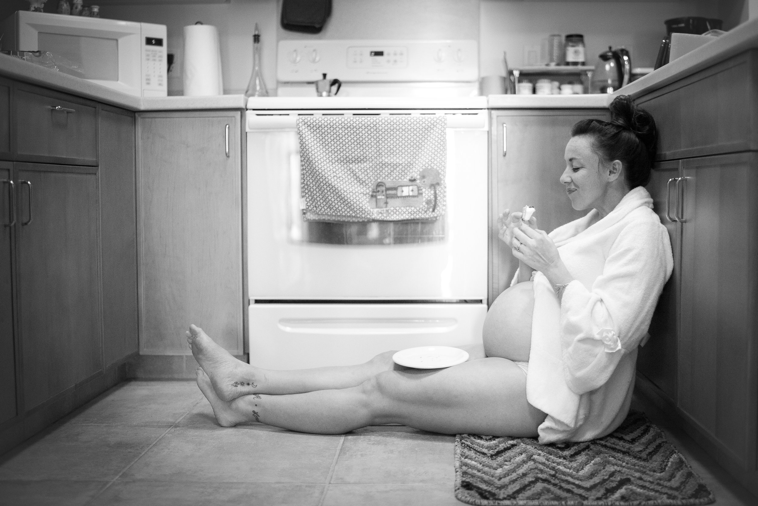 A woman sitting on the kitchen floor with her back against the cabinets, smiling and holding food. She has a mat underneath her feet, and a plate on her belly. The kitchen has appliances like a microwave, stove, and various items on the countertops. 