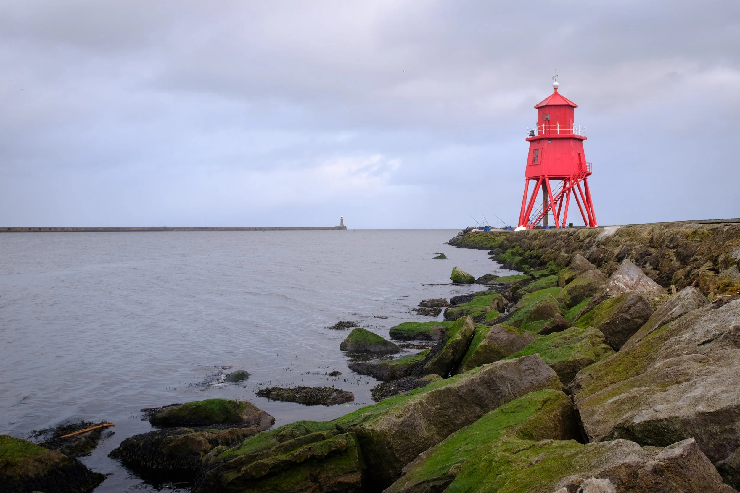 Red lighthouse on rocky green and brown shoreline with water and cloudy sky in the background.