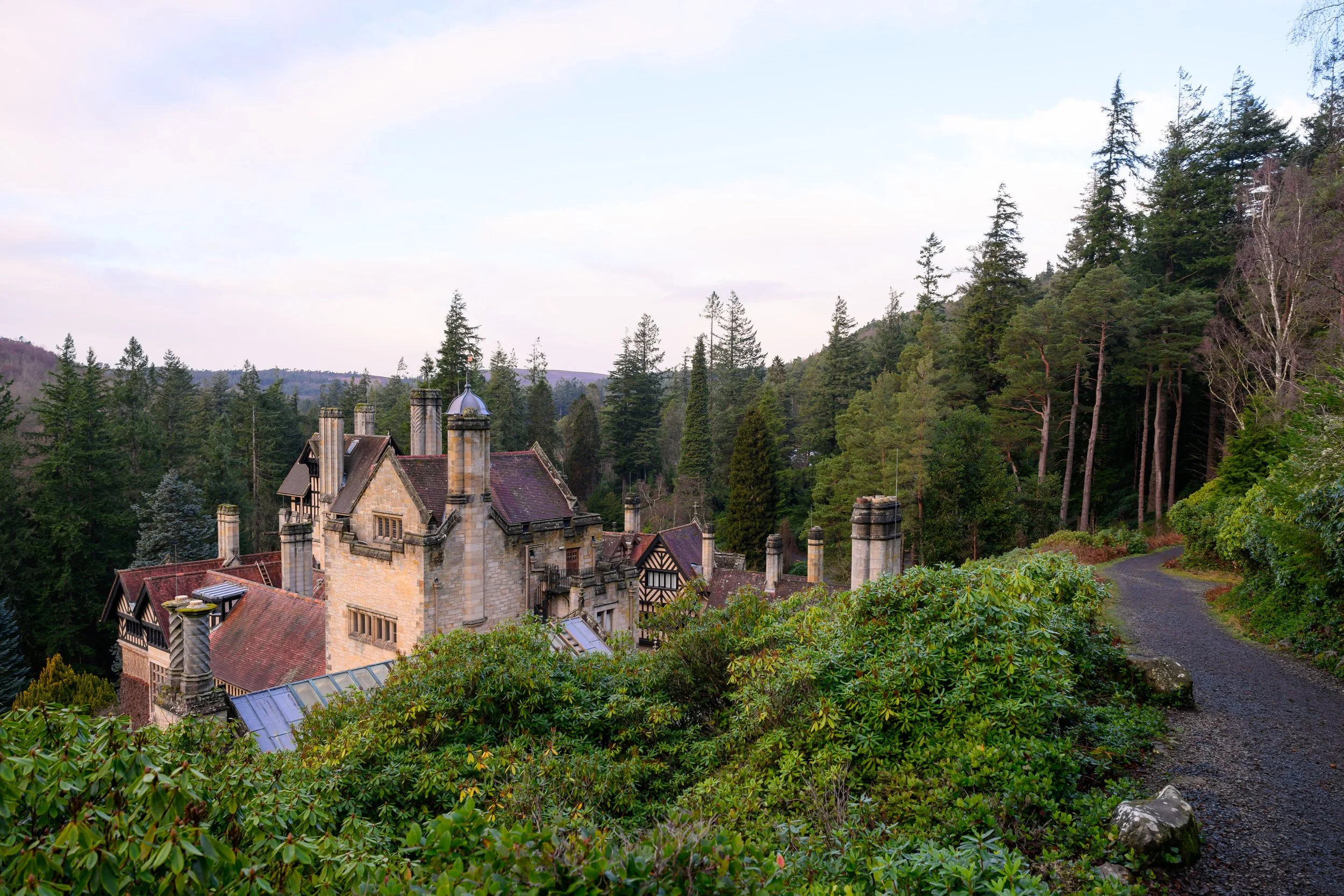 A historic stone mansion with multiple chimneys and steep roofs, surrounded by dense green trees and a winding gravel path in a forested landscape.