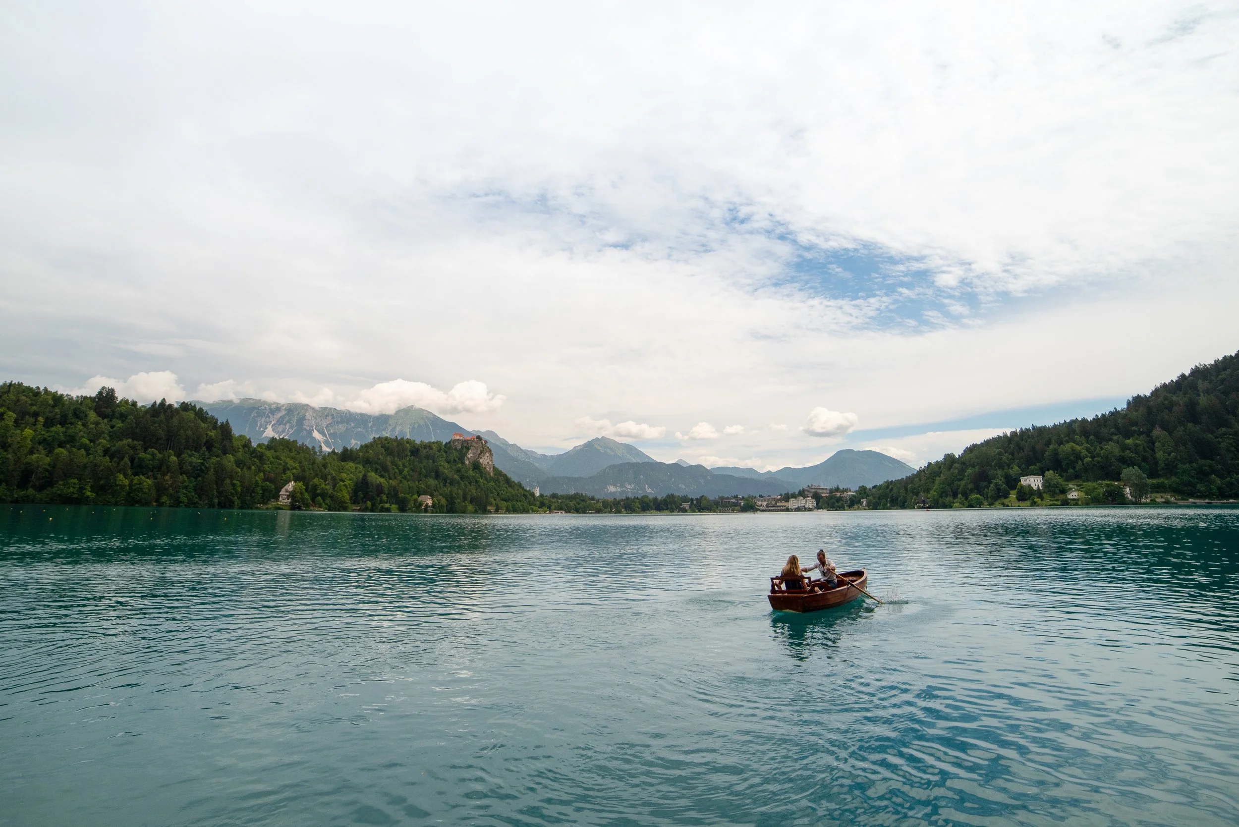 Two people in a small wooden boat on a calm lake surrounded by lush green hills and mountains under a partly cloudy sky.
