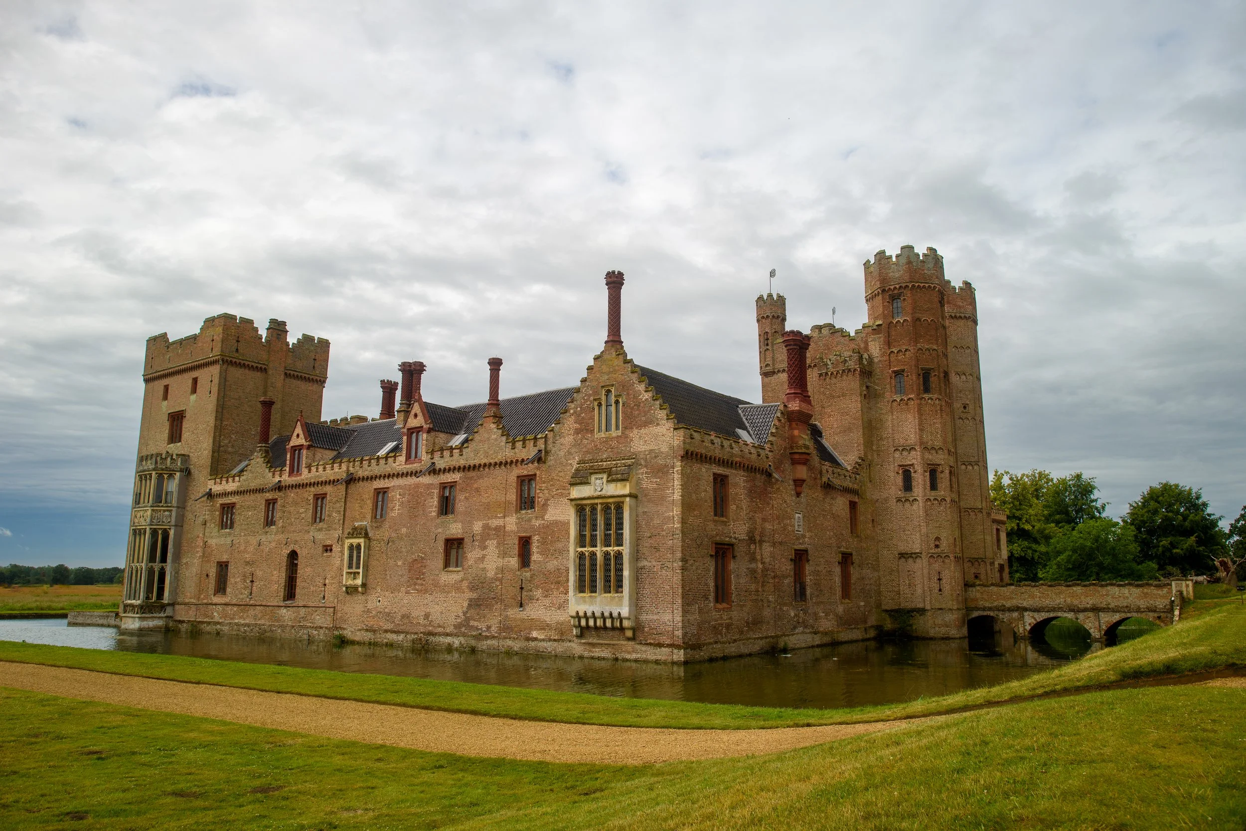 A historic brick castle with multiple towers surrounded by a moat, under a cloudy sky.