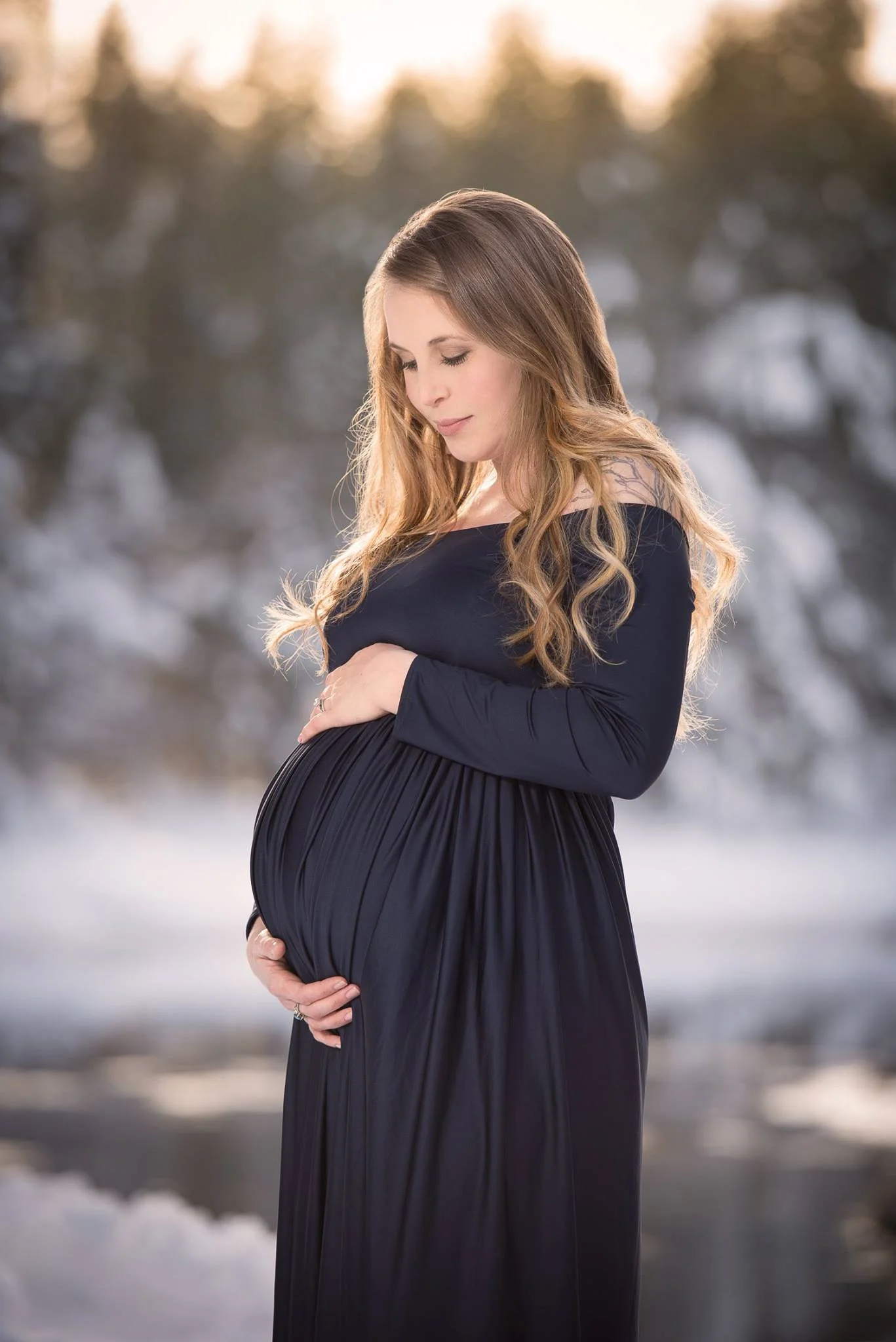A pregnant woman with long, wavy hair in a black dress looking down at her belly outdoors during winter.