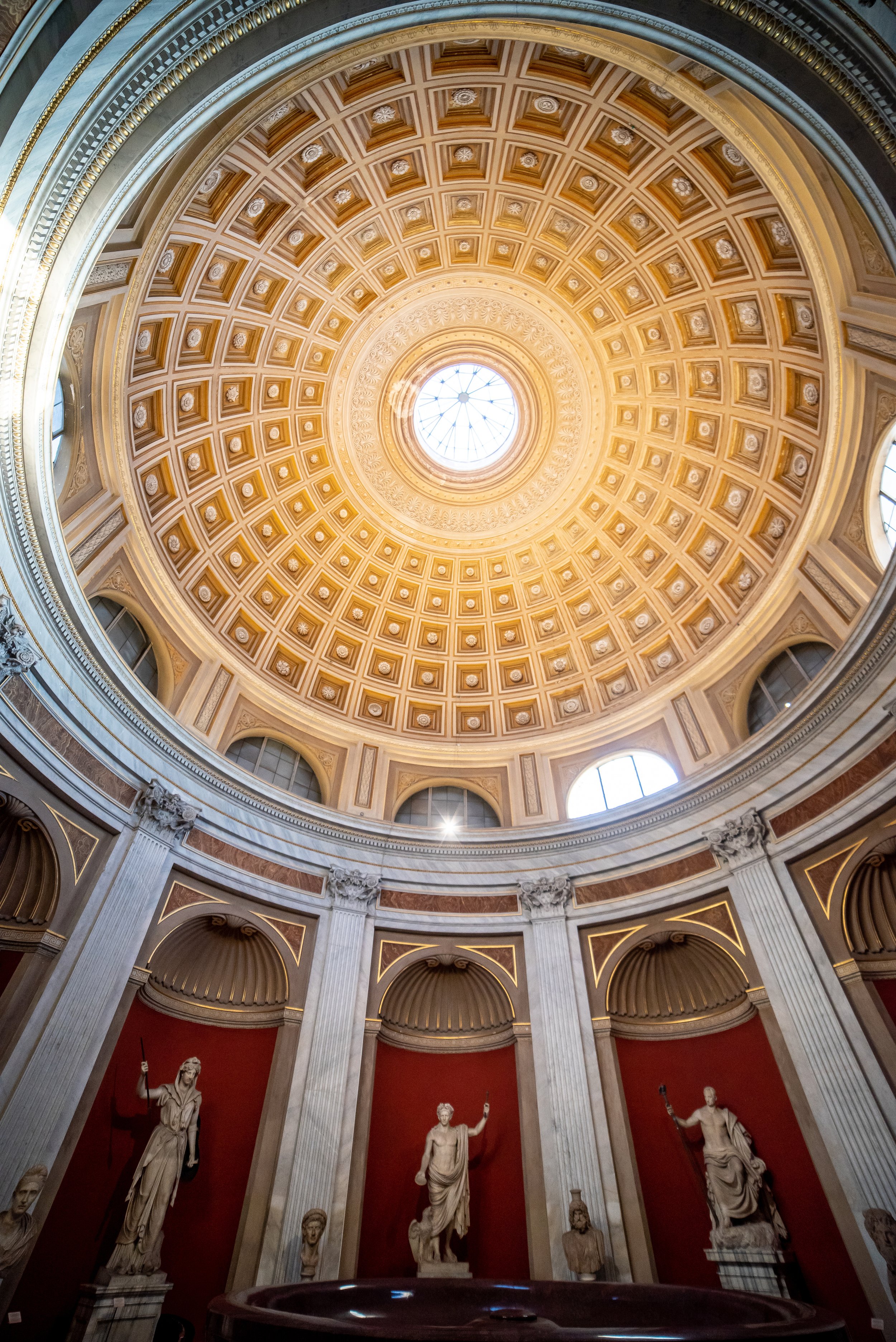 Interior view of a grand rotunda with a detailed golden coffered ceiling and a central skylight, displaying classical statues against red walls.