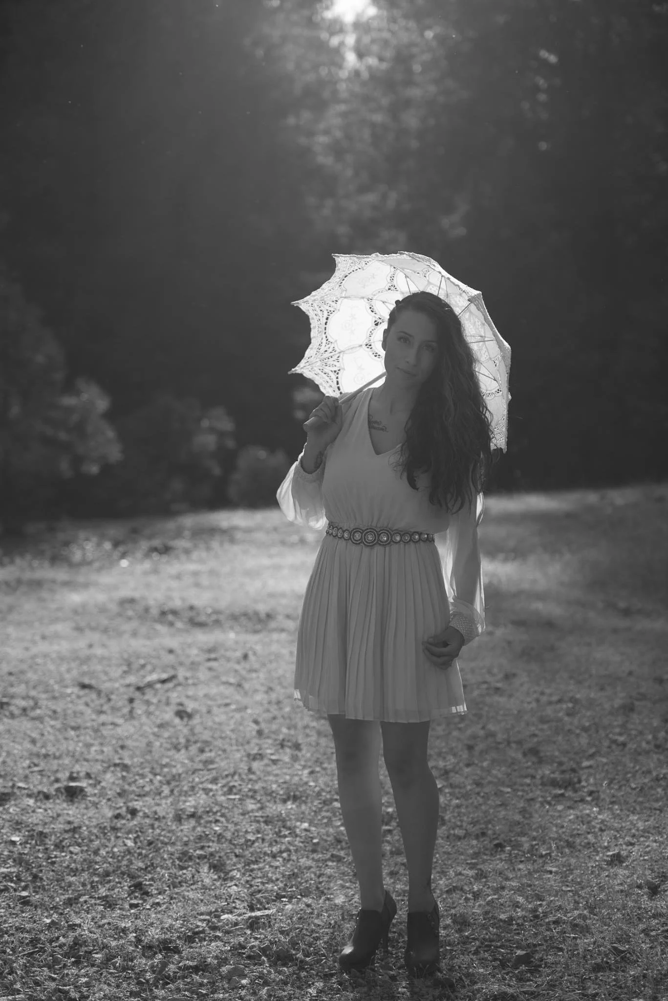 A woman with curly hair standing outdoors at sunset, holding a lace umbrella, wearing a pleated skirt and high heels.