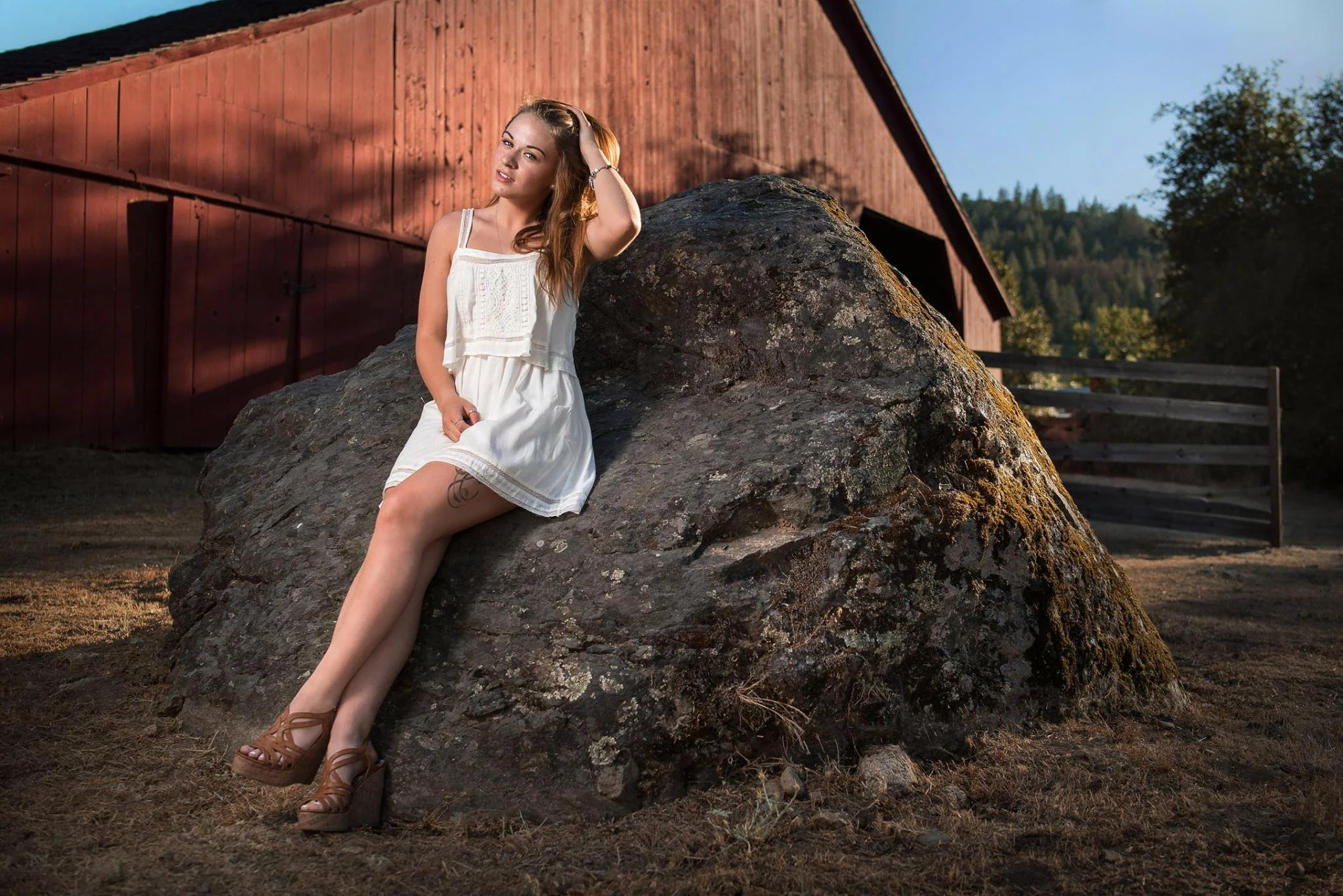 A woman in a white dress and brown wedge sandals sitting on a large rock in front of a red barn with trees in the background.