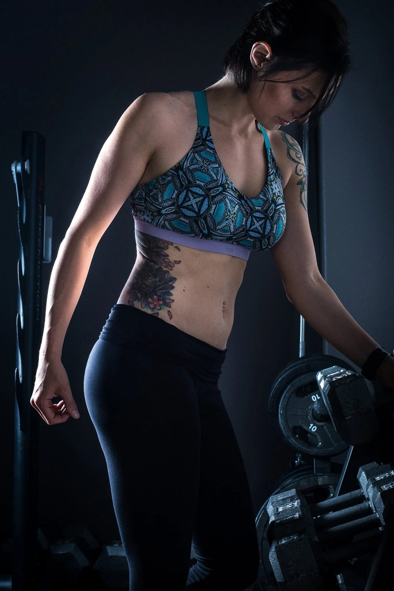A woman with short dark hair wearing a colorful sports bra and black leggings, exercising with dumbbells in a dark gym.