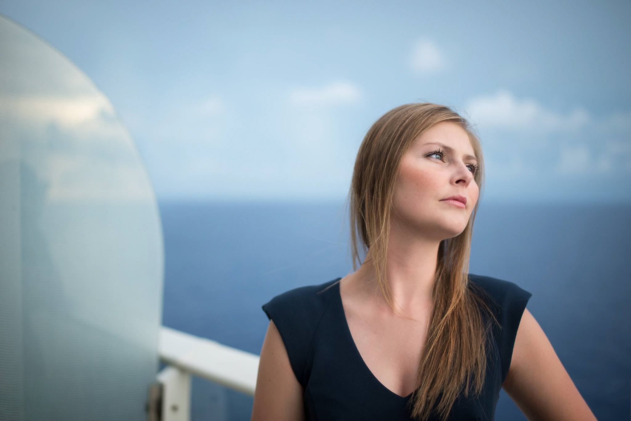A woman with long blonde hair looking thoughtfully toward the sky, standing on a boat or ship with water in the background.